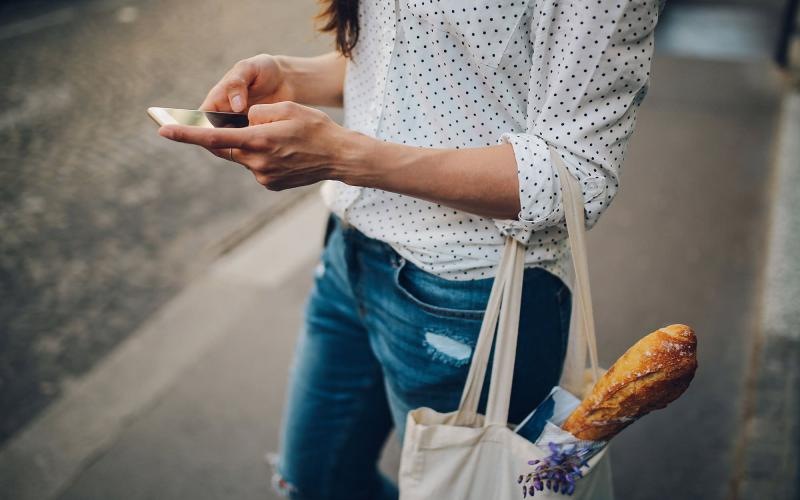 Close to the center of everything woman walks down the street and texts with a bag of groceries on her arm